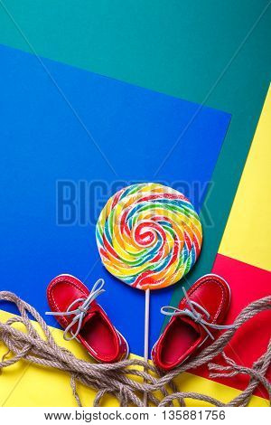 Small red boat shoes near big multi-colored lollipop and rope on colored background. Top view. Copy space frame