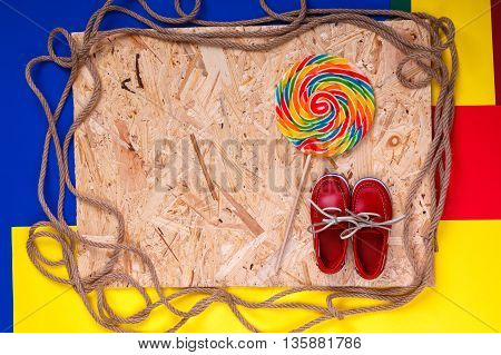 Small red boat shoes near big multi-colored lollipop and rope on wooden desk with colored background. Top view Frame. copy spase.