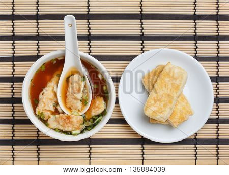 Bowl of delicious traditional japanese miso soup with wakame seaweed and white ceramic spoon in it plate with sliced fried tofu standing on striped bamboo mat.