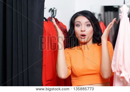 Young woman astonished in a fitting room looking at the camera. Fashionable woman shopping in a clothing store. Woman shopper in a fitting room. Woman shopping for dress. Woman shopaholic. Shopping
