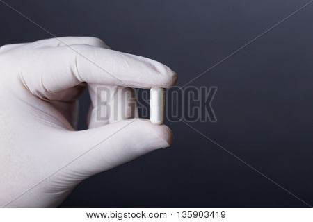 Hand in white latex glove holding white capsule. Doctor or nurse showing medicine. Dark background