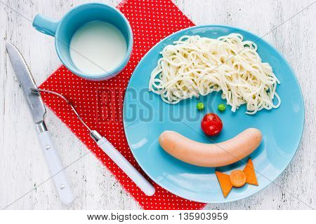 Dinner or breakfast for kids - spaghetti with sausage and vegetables in the shape of clown face. Creative food art idea for children meal top view