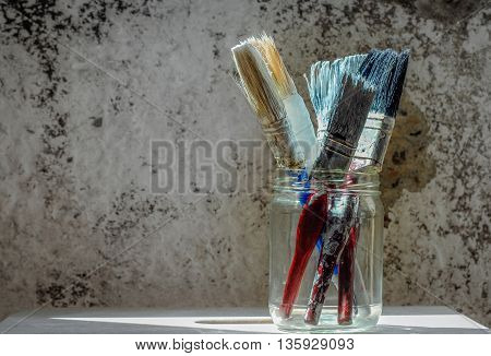 Old paint brushes in a glass jar on grungy wall background