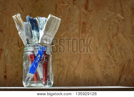 Old paint brushes in a glass jar on wooden panel background