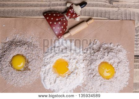 Three eggs broken on heaps of white and whole wheat flour bran with red dress porcelain figurine toy culinary utensils rolling pin on timber background