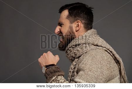 Unleashing his emotions. Side view of furious young bearded man shouting while standing against grey background