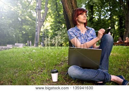 Beautiful young freelancer woman using laptop sitting under the tree in the park.Happy smiling girl working online.Studying and learning using notebook computer.Freelance workbusiness people concept.