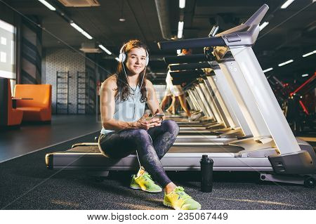 Beautiful Young Caucasian Girl Sportswoman Sitting, Resting After Training On Treadmill Against The 