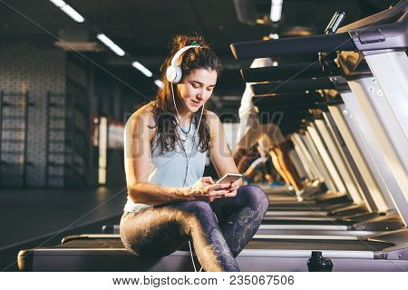 Beautiful Young Caucasian Girl Sportswoman Sitting, Resting After Training On Treadmill Against The 