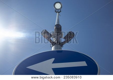 Three Surveillance Camera Attached On Led Light Pole With Direction Sign. Blue Sky With Sunray As Ba
