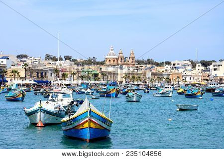 Marsaxlokk, Malta - April 1, 2017 - Traditional Maltese Dghajsa Fishing Boats In The Harbour With Th