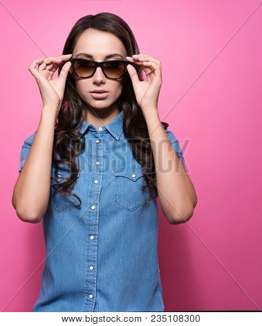 Attractive Young Woman With A Neutral Expression Holds For Glasses. Stylish Look, Blue Denim Shirt. 
