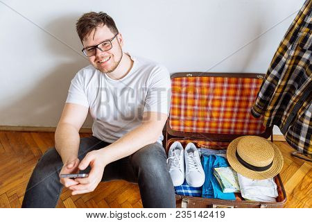 Young Man Sit On Floor Checking List On Mobile. Valise Packing For Trip.travel Concept. Copy Space.