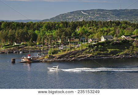 Oslofjord Bay With Houses On A Rock, Moving Speedboat And Holenkollenem In The Background.