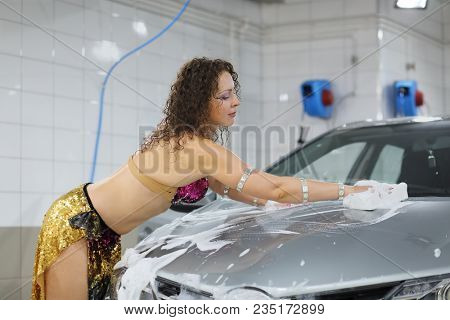 Beautiful woman in a shiny dress with a foam sponge washes the hood of a silver car on a car wash