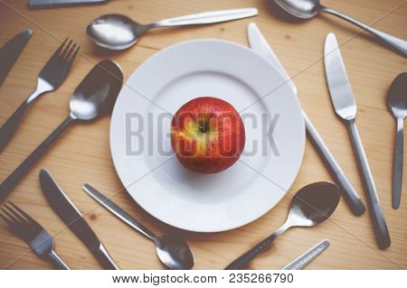 Still Life: An Red Apple Lying On Small Plate. Spoons, Tea Spoons, Forks And Knives On The Wooden De