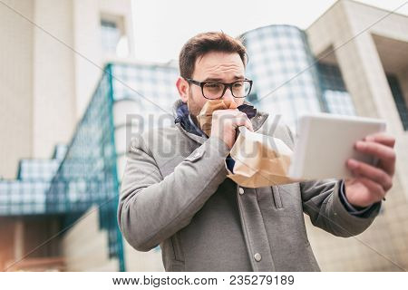 Businessman Holding Paper Bag Over Mouth As If Having A Panic Attack And Looking In Digital Tablet.