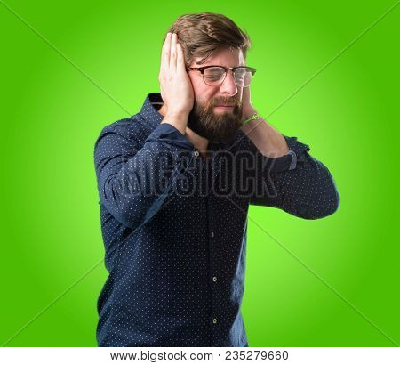 Young hipster man with big beard covering ears ignoring annoying loud noise, plugs ears to avoid hearing sound. Noisy music is a problem. over green background