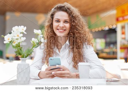 Photo Of Attractive Curly Woman With Smile, Dressed In Elegant Clothing, Reads Pleasant Sms Message,