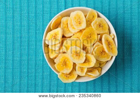 Heap Of Dried Banana Chips Snack In White Bowl On Blue Turquoise Mat On Table