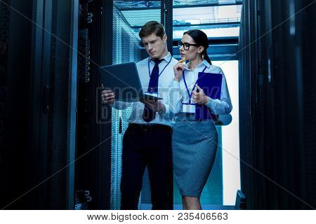 Our Project. Serious Professional Co-workers Standing Near A Service Cabinet And Discussing Work