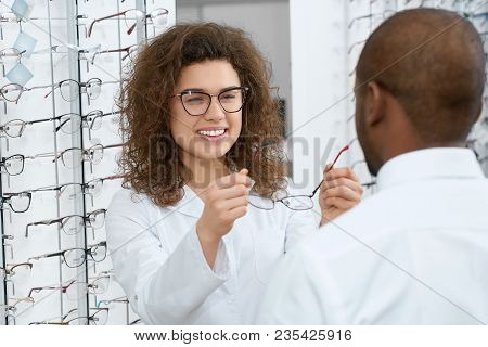 Backview Of Man Trying On Eyeglasses In Optical Shop. Young Doctor Is Helping The Client To Choose T