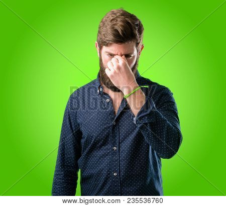 Young hipster man with big beard with sleepy expression, being overworked and tired, rubbes nose because of weariness over green background