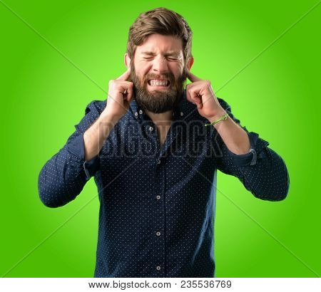 Young hipster man with big beard covering ears ignoring annoying loud noise, plugs ears to avoid hearing sound. Noisy music is a problem. over green background