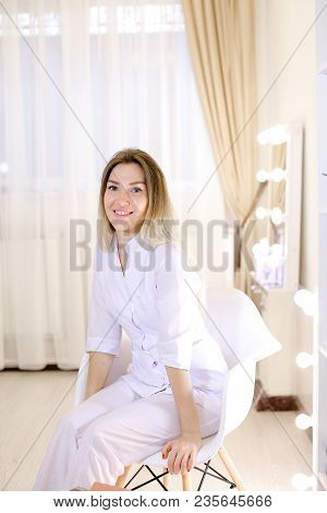 Blonde Nurse Sitting In Chair At Cabinet And Wearing White Suit. Concept Of Beauty Salon And Cosmeto