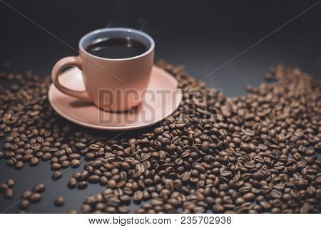 Cup Of Black Steaming Coffee On A Spilled Roasted Coffee Beans On A Black Background.