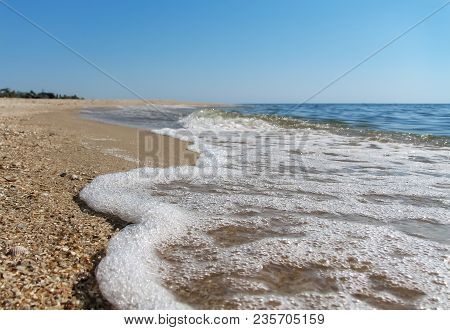 Sea Foam On The Shore From Seashells And Sand On A Background Of A Calm Sea And Clear Blue Sky