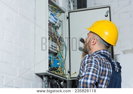 Professional Electrician With Flashlight Working By Distributor Box