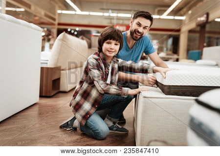Bearded Man With Young Son Is Testing Mattress For Softness. Testing Softness Of Mattress. Choosing 