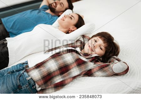 Young Beautiful Couple With Cute Little Boy Lies On Bed In Mattress Store. Family Checking Softness 