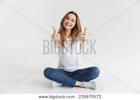 Happy woman in t-shirt sitting on the floor and showing thumbs up while looking at the camera over gey background