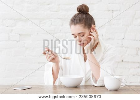 Portrait Of Young Bored Woman Eating Cereals With Bad Taste In Her White Loft Kitchen, Sitting At Wo