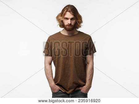 Young European Masculine Bearded Man Standing With Hands In Pockets, In Brown Tshirt, Isolated On Gr