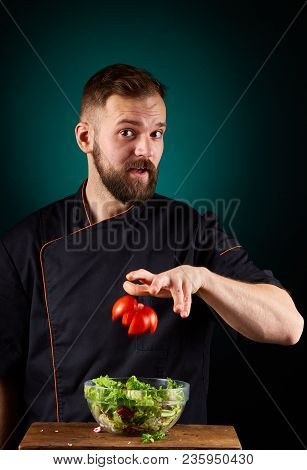 Portrait Of A Handsome Mischevious Male Chef Cook In Black Uniform Making Delicious Vegetable Salad 