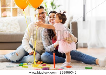 family, holidays and people concept - happy mother, father and little daughter reaching to balloons at home birthday party
