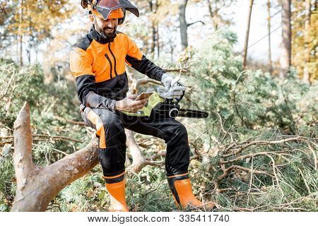 Portrait Of A Professional Lumberman In Protective Workwear Sitting With Mobile Phone On The Felled 