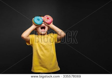 Happy cute boy is having fun played with donuts on black background wall. Bright photo of a boy. Colored donuts