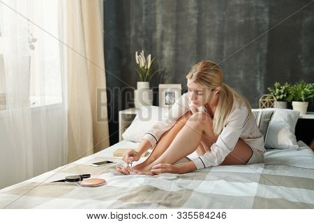 Young blond woman in silk pajamas sitting on bed and painting toenails with nail polish in the morning