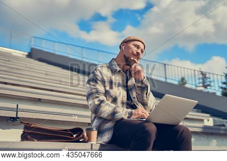 Thoughtful Young Bearded Man With Mustache Works Outisde While Holds Laptop On Legs. City Space