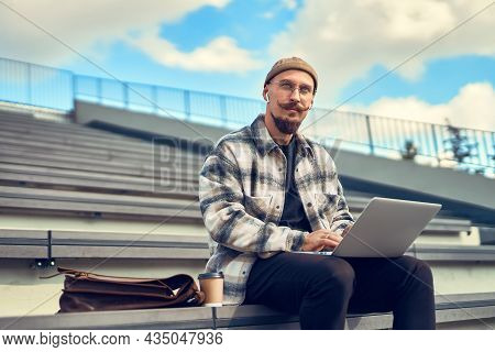 Thoughtful Young Bearded Man With Mustache Works Outisde While Holds Laptop On Legs. Look Sideways