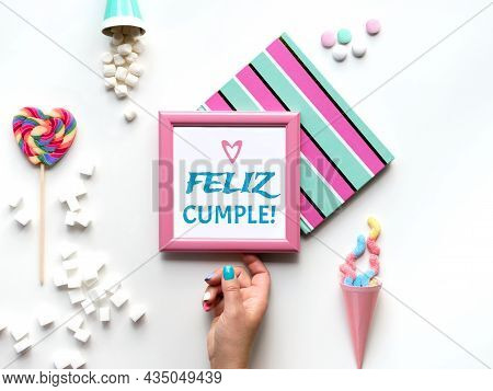 Text Feliz Cumpleanos Means Happy Birthday In Spanish. Pink Picture Frame In Hand. Sweets And Party 