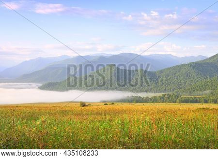 Fog Over A Green Meadow Against The Background Of Blue Mountains Under The Morning Sky. Siberia, Rus