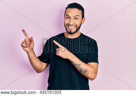 Hispanic man with beard wearing casual black t shirt smiling and looking at the camera pointing with two hands and fingers to the side. 