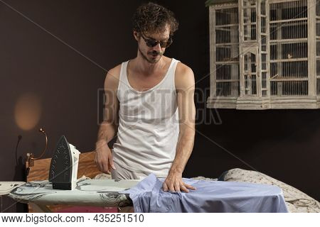 Young man alone in his home ironing a shirt in a white tank top and in his underwear.