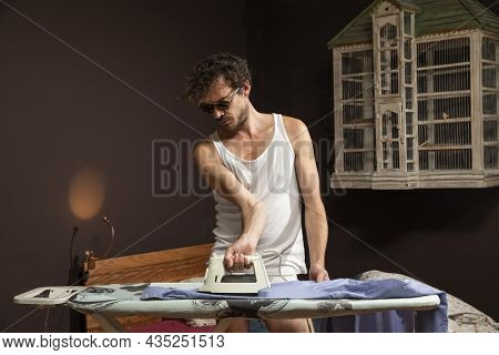 Young man alone in his home ironing a shirt in a white tank top and in his underwear.