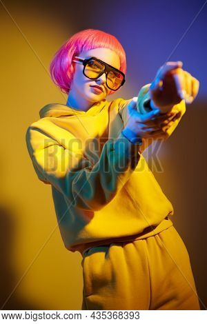 Modern girl with bright pink hair poses in yellow sunglasses and yellow hoodie suit in mixed color light pointing her finger. Sport chic fashion. Youth fashion and beauty trends. 